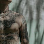 Detailed close-up of a tattooed male torso with varied designs, captured in soft shadowed lighting.